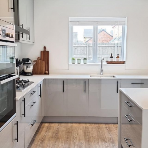 Gloss grey Woodbury kitchen shown with white worktops and black skinny handles. wooden and matt black accessories are also shown 