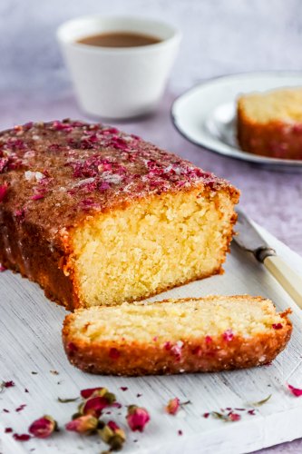 Lemon and Rose Drizzle Cake laid on a chopping board surrounded by dried rose petals. There is a knife on the chopping board and a cup of tea to the side of the board