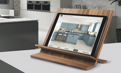 Ipad is shown in a kitchen on a wooden tablet stand with a white kitchen in the background. Screen shows the Symphony dream kitchen visualiser with a blue kitchen featured
